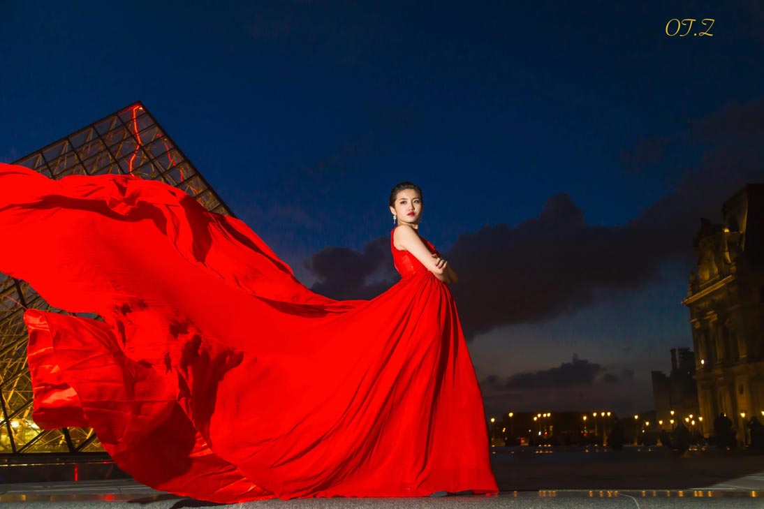 Woman in a flowing red gown beside the Louvre Pyramid at dusk